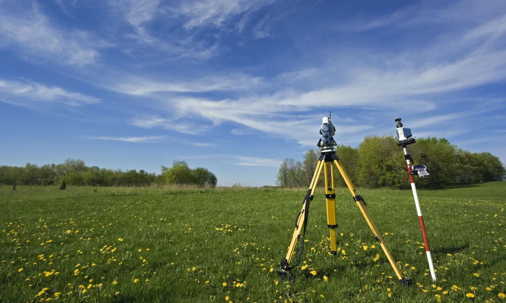 Survey equipment in a grassy field with dandelions sprinkled throughout, a blue sky, and some trees in the distance.