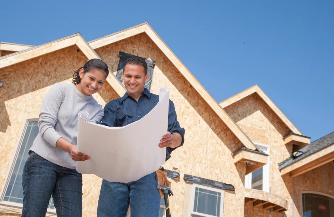 A male and female couple looking at house plans with a partially constructed home behind them.