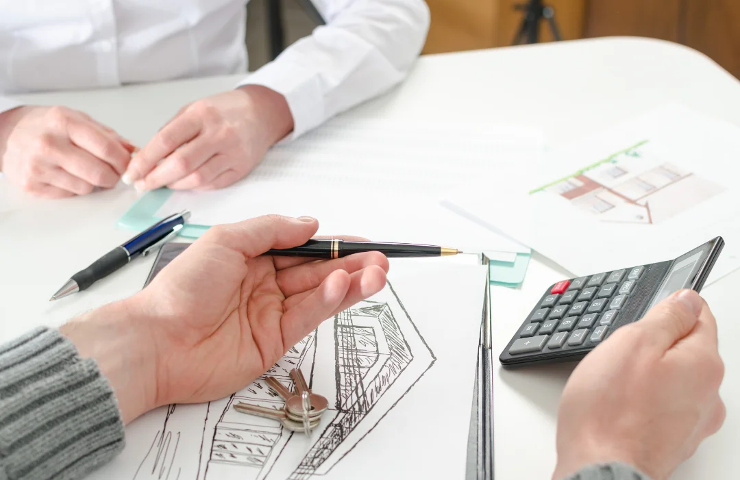 A mortgage broker's hand holding a calculator, showing it to a young woman who is sitting opposite. Broker is holding a pen, using it to point to a figure on the screen. There are house plans and keys on the desk.