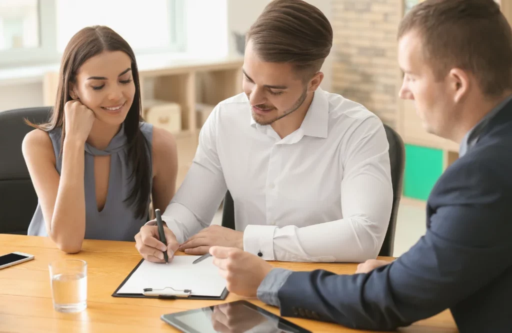 A young man and a woman signing mortgage papers with a local mortgage broker helping them along.