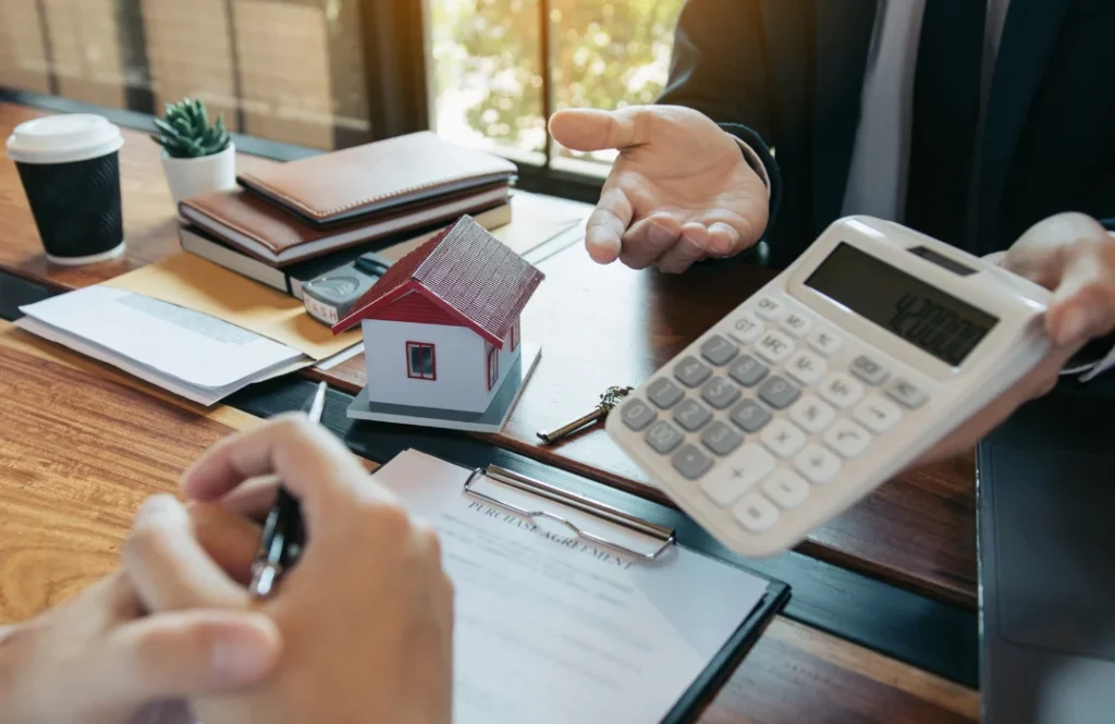 Mortgage broker holding a calculator with 420,000 dollars on the screen, showing it to client across the desk with expressive hands. There's a little house on the desk and a purchase agreement on a clipboard.