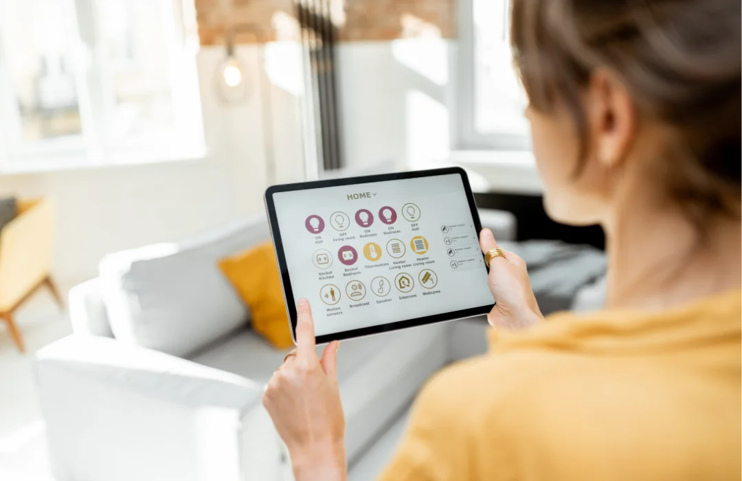 CMS-Smart-Home-Tech-2 A woman looking at a tablet with smart home icons on the screen while standing in her living room.