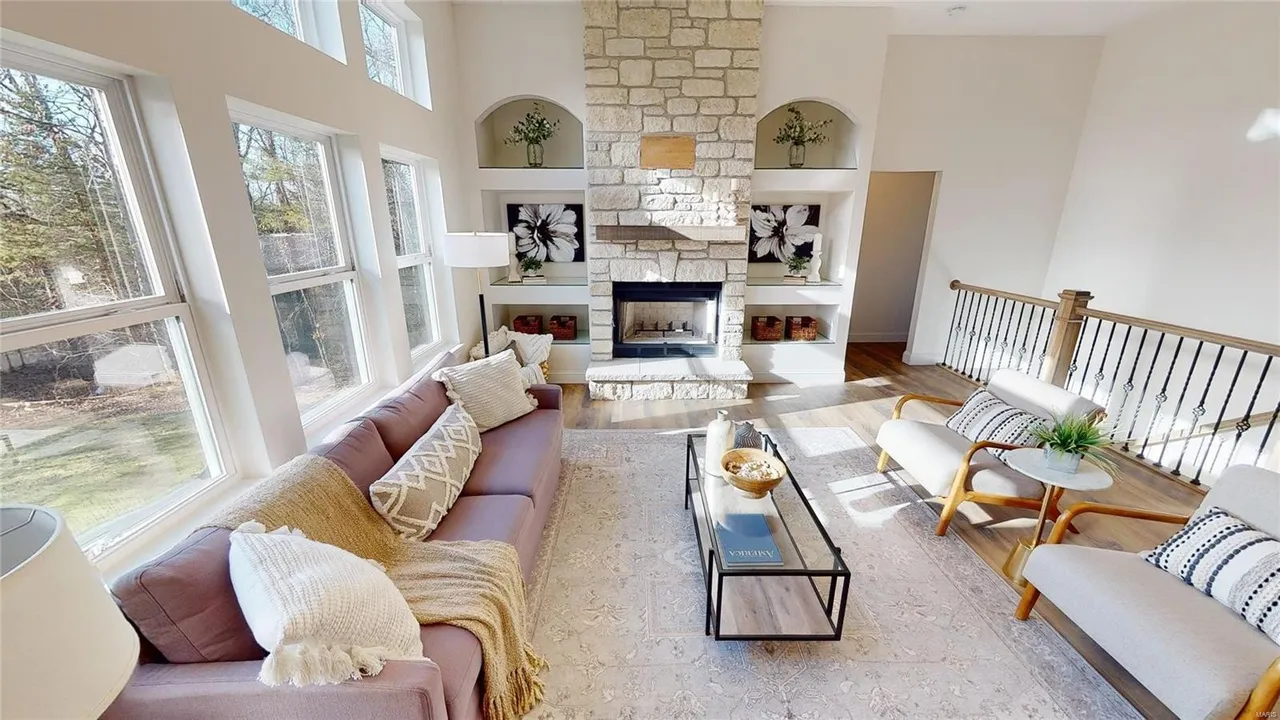 A new-home living room with a stone fireplace and shelving next to the fireplace. Earth tones, large windows, and hardwood floors. Very cozy open plan.